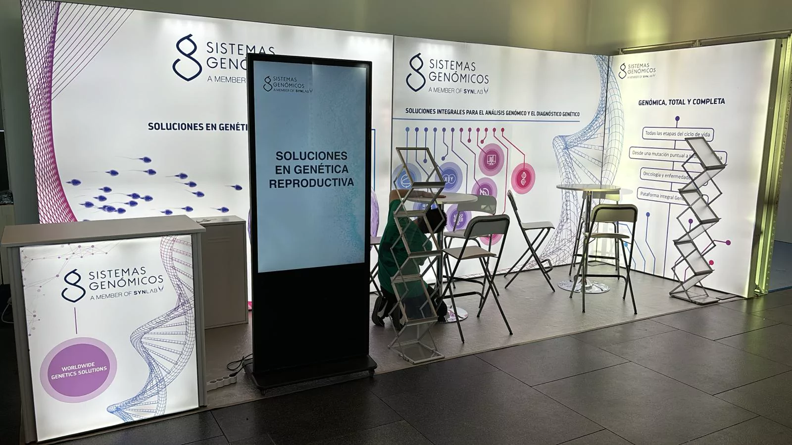 Stand de Sistemas Genómicos con paredes retroiluminadas, tótem digital y mostrador